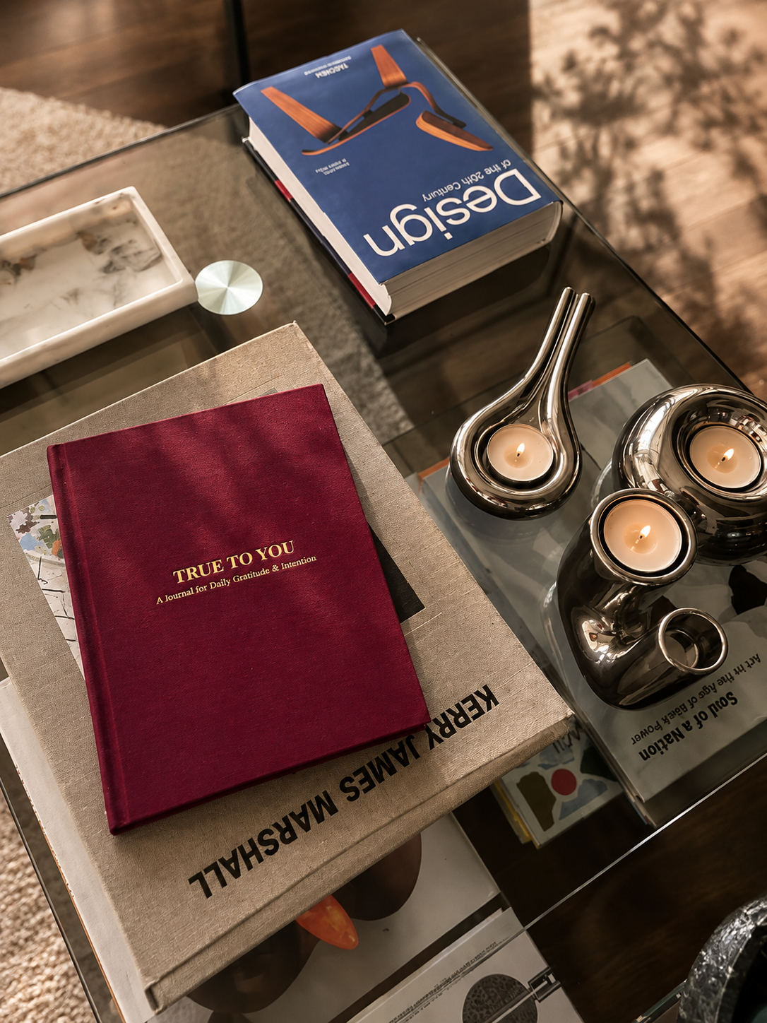A bordeaux True To You journal resting on art books on a glass coffee table beside lit candles